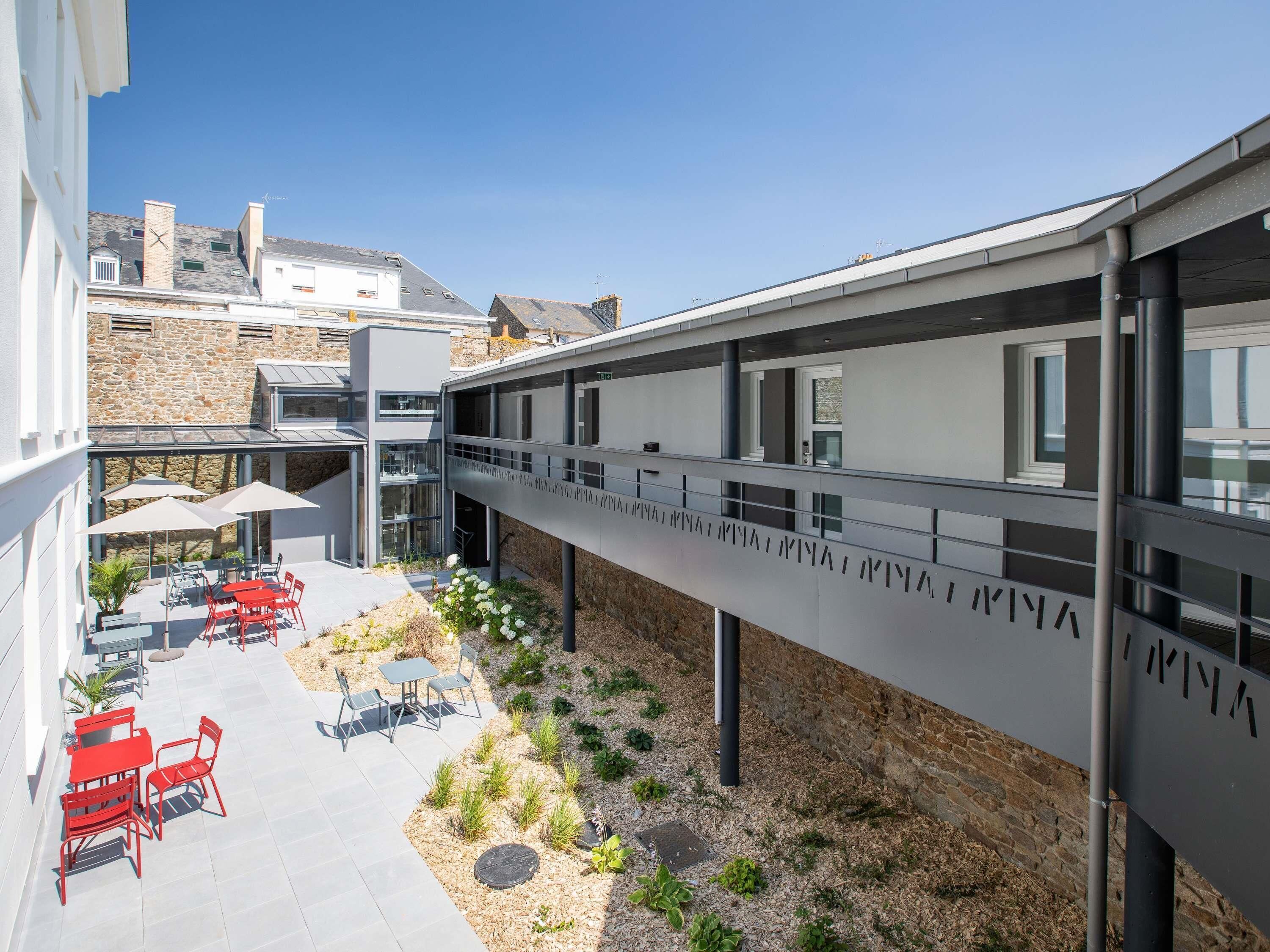 A modern building with a covered outdoor area featuring red chairs and tables, a stone wall, and a bridge with text.