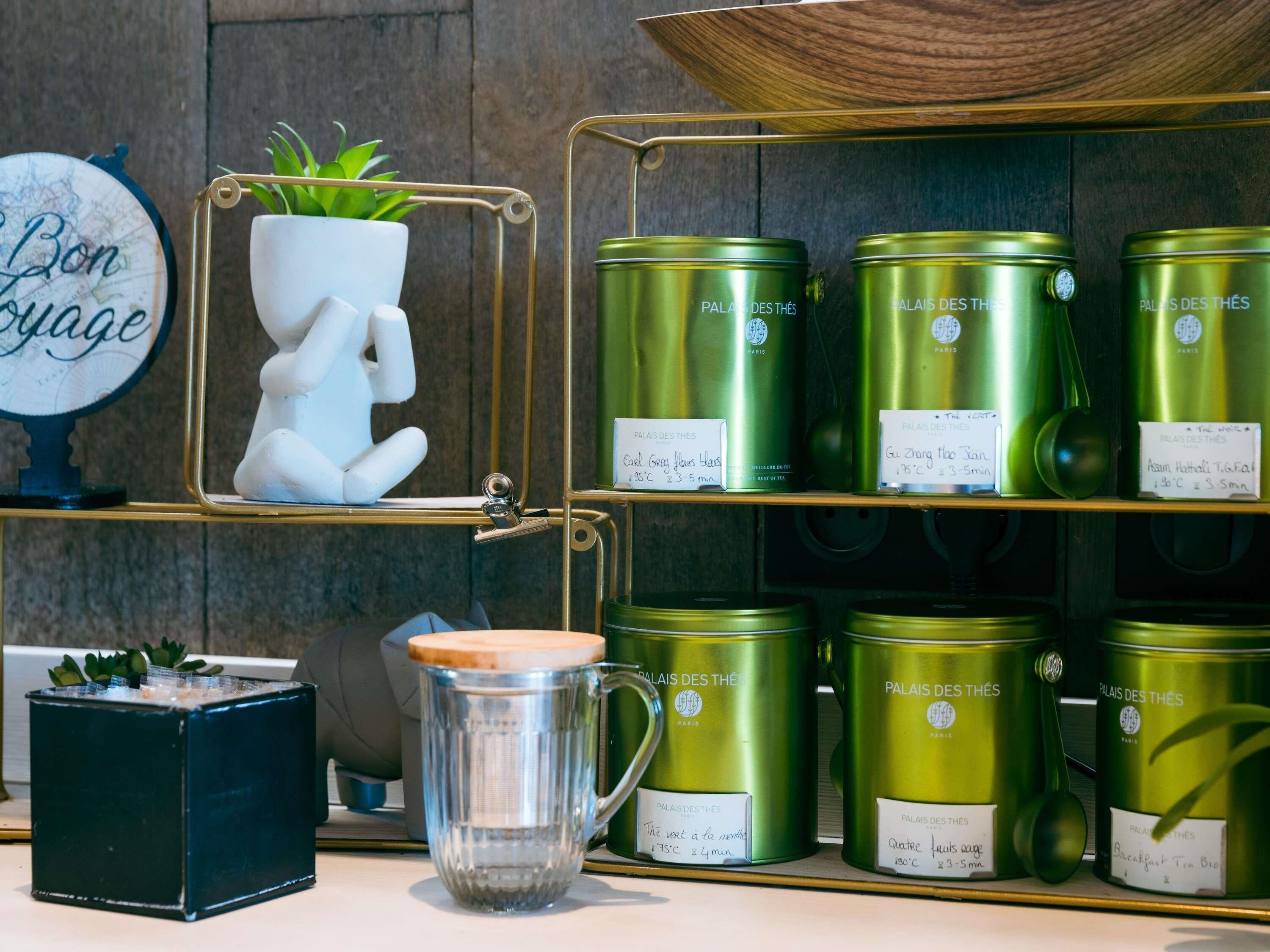 Una estantería con recipientes verdes etiquetados 'Palais des Thés Paris' y una taza de vidrio transparente con tapa de madera.