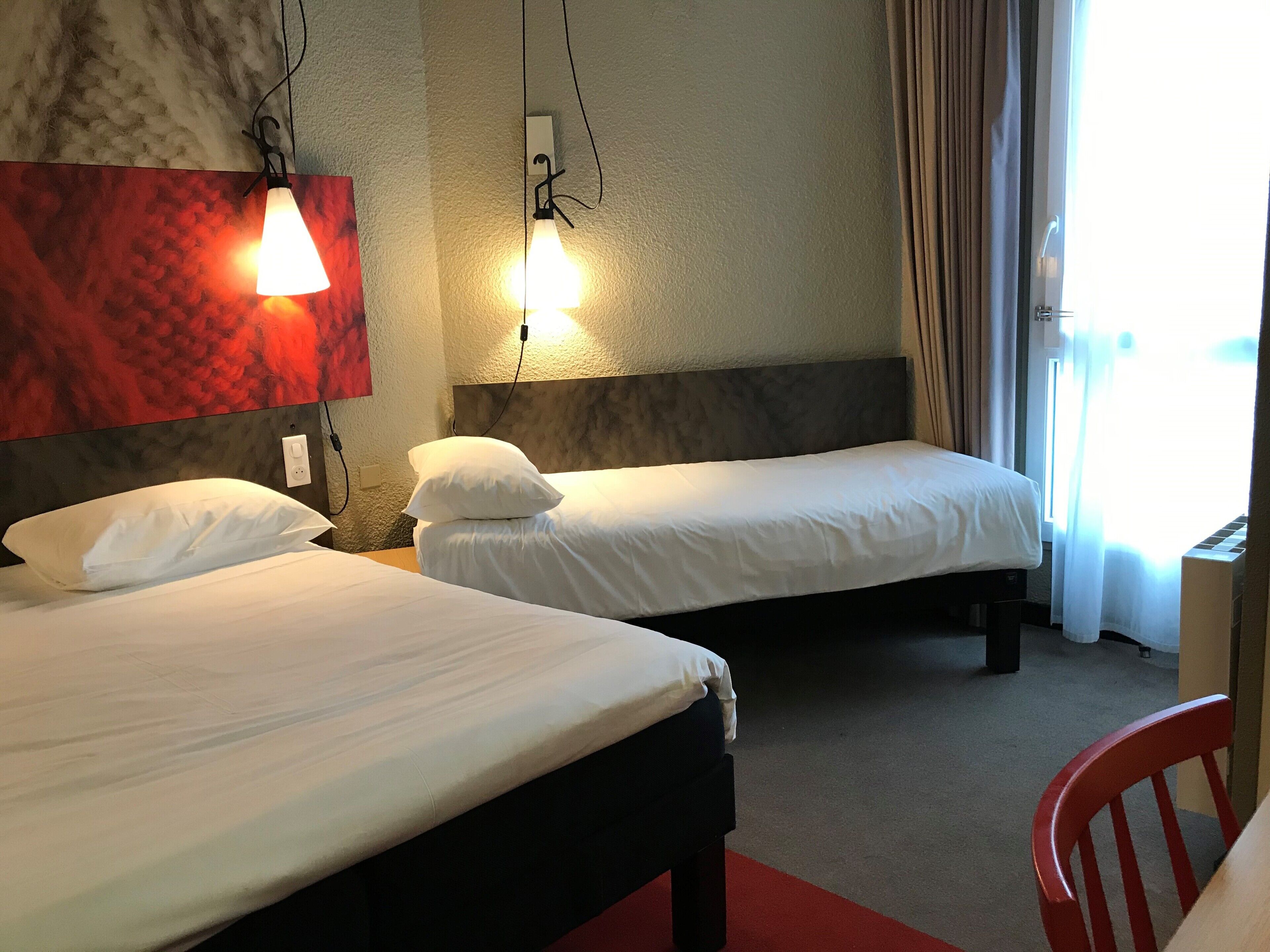 Una habitación de hotel con dos camas, una alfombra roja, un sillón rojo y una ventana con cortinas.