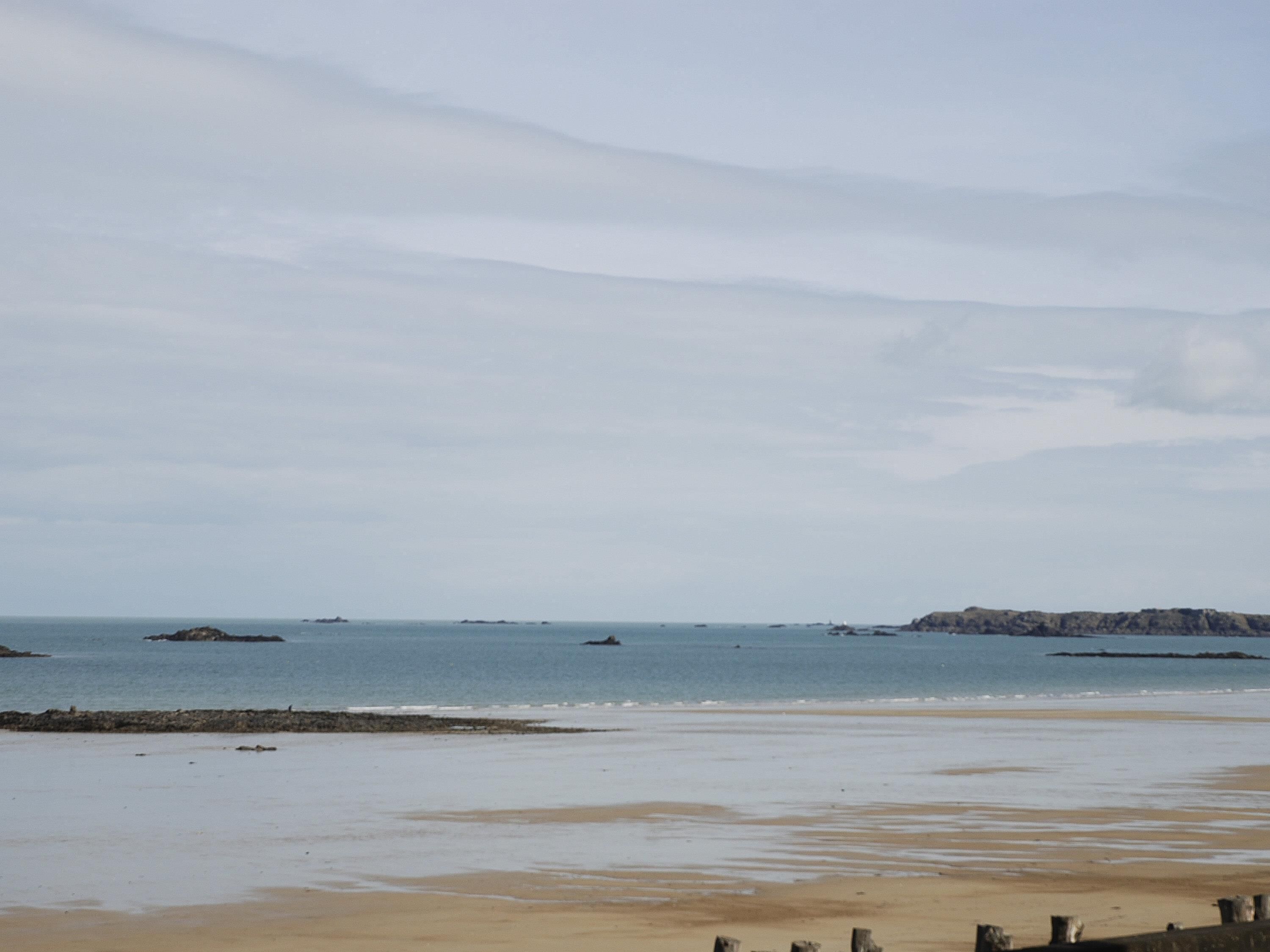 A beach with calm waters, distant rocky outcrops, and a clear sky.