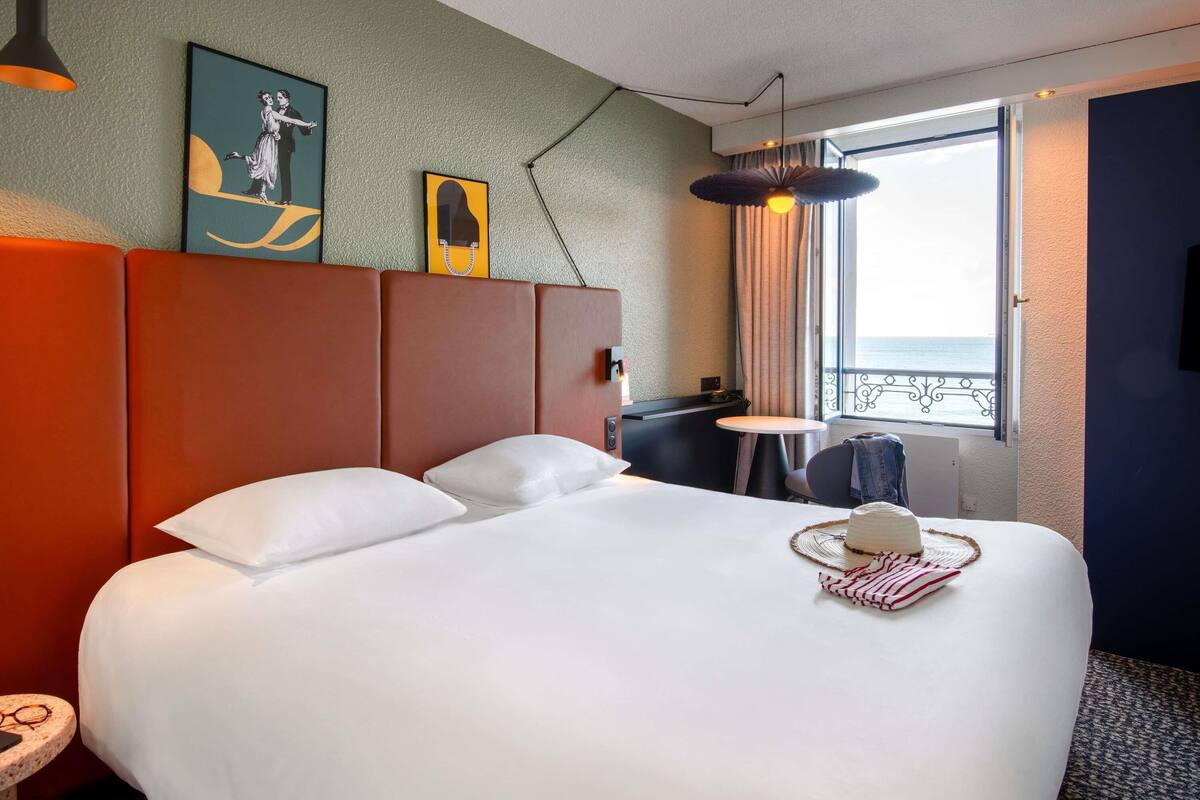 Une chambre d’hôtel avec un grand lit, une petite table, une chaise, une fenêtre donnant sur la mer et des œuvres d’art accrochées au mur.