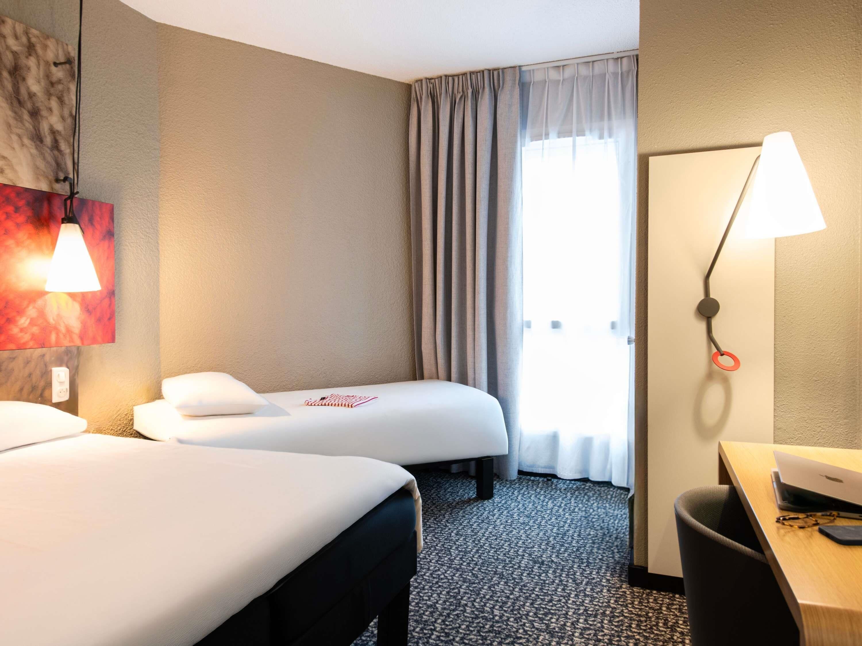 A hotel room with two beds, a desk, a lamp, a window with curtains, and a wall-mounted artwork.