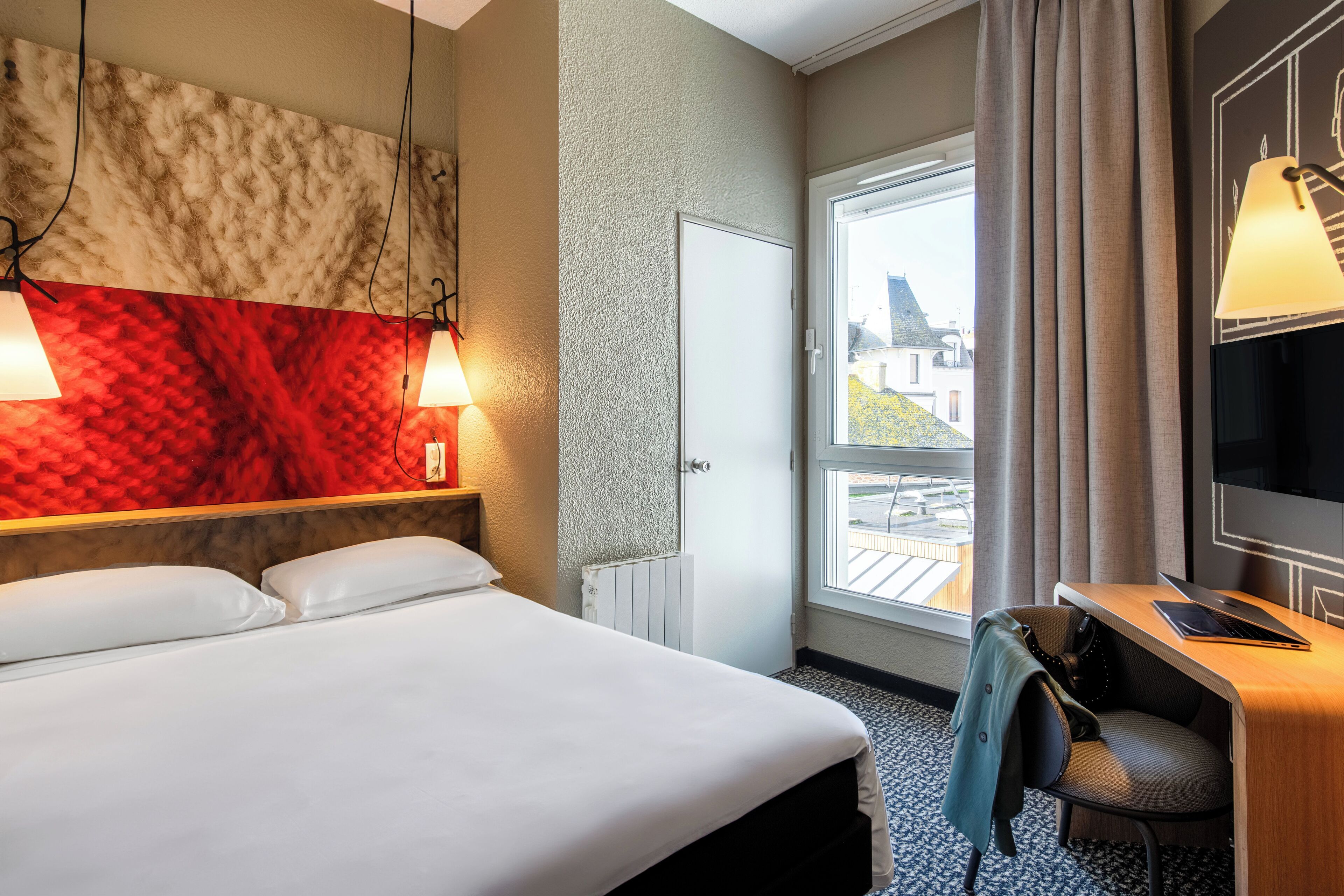 Een hotelkamer met een bed, een bureau met een laptop, een televisie en een raam met uitzicht op gebouwen.