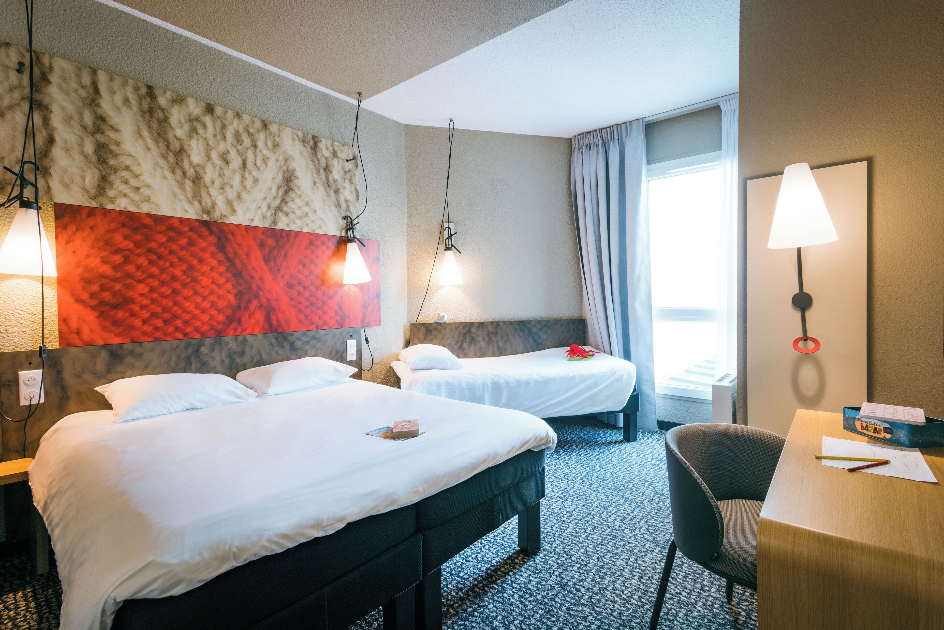 Een hotelkamer met twee bedden, een bureau en een stoel. Er zijn twee wandlampen en een raam met gordijnen.