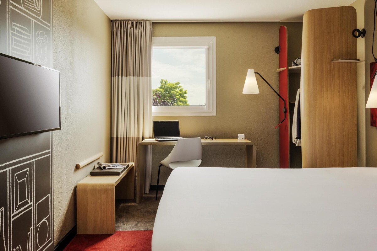 A hotel room with a bed, desk, chair, TV, and a window with curtains.