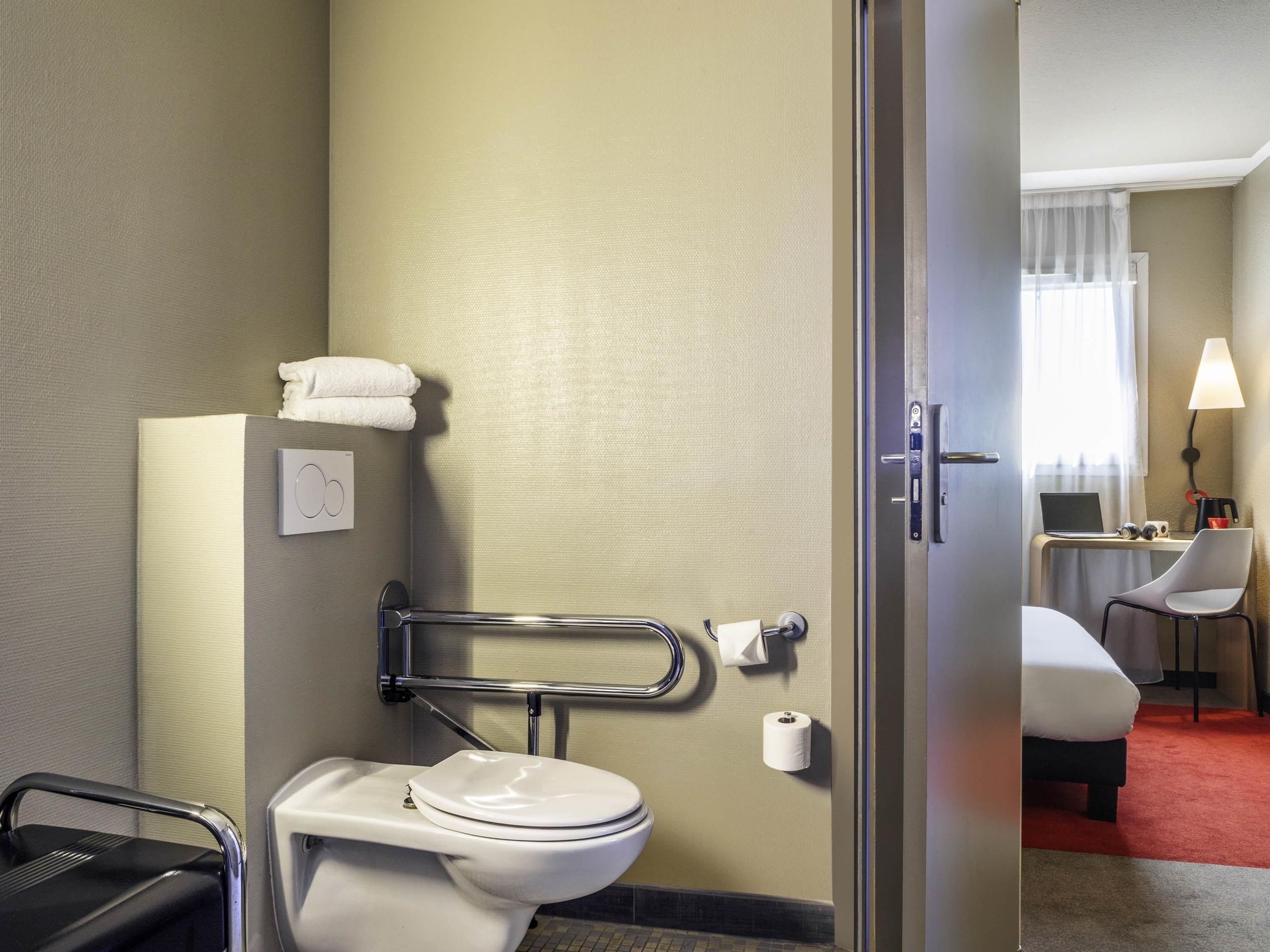 Ett hotellbadrum med toalett, stödhandtag, handduksställ och en synlig dörr som leder till ett sovrum.