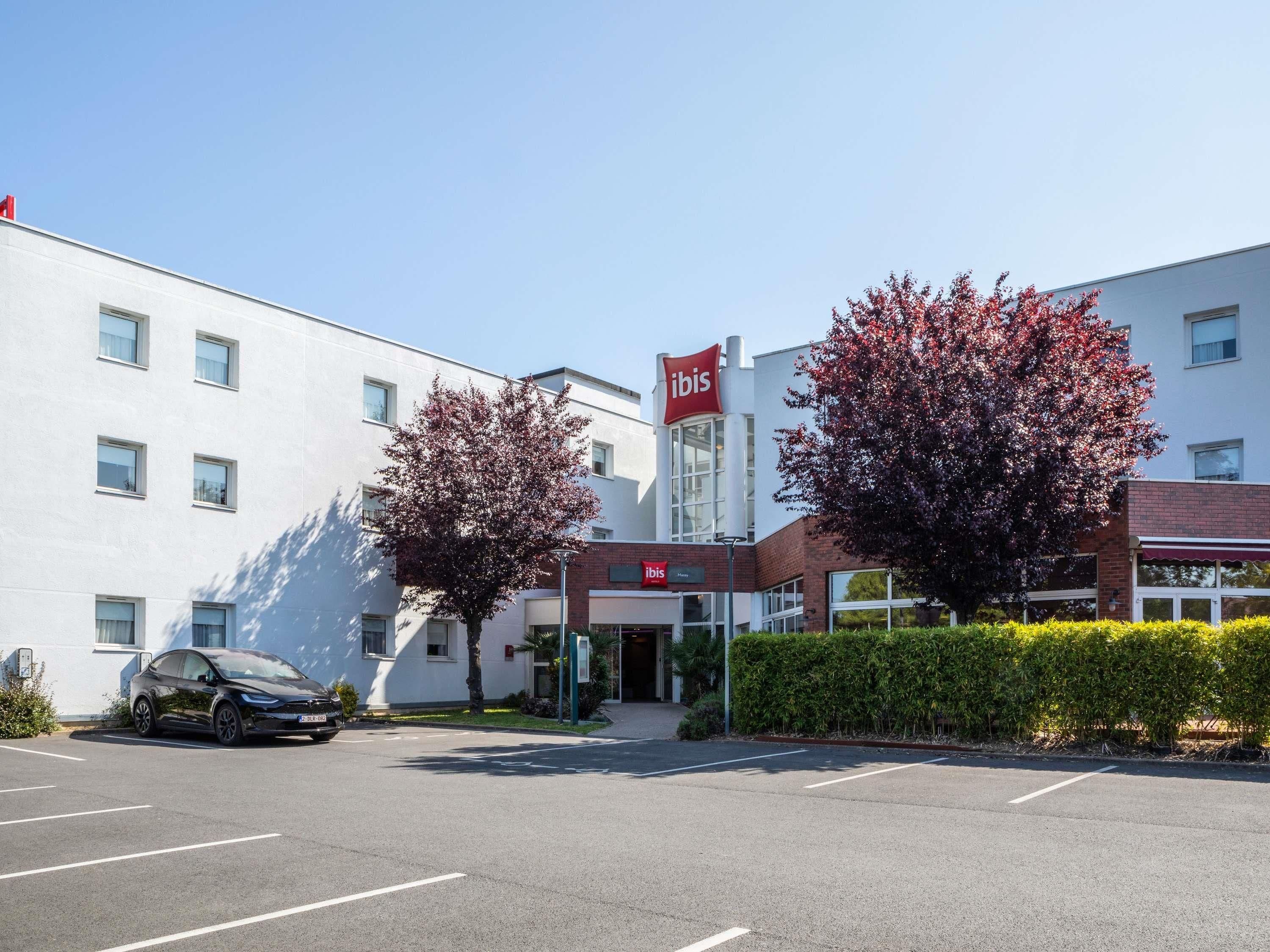 A modern hotel with a prominent 'ibis' sign, a parking lot, and two trees with red foliage.