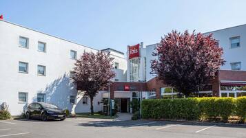 Um hotel moderno com uma placa grande da 'ibis', um estacionamento e duas árvores com folhagem avermelhada.