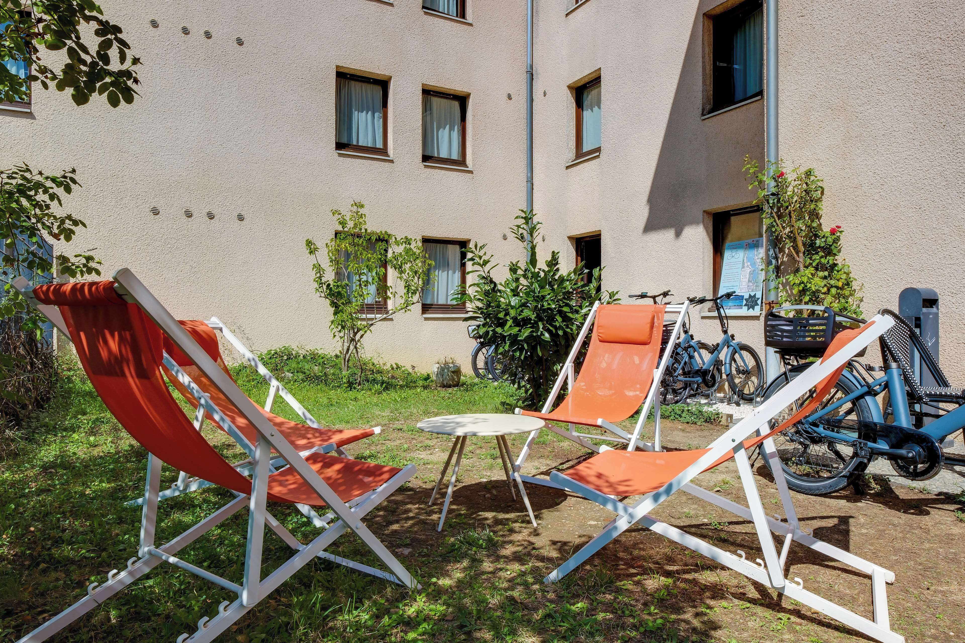 Une cour avec deux fauteuils de jardin orange, une petite table ronde et des vélos appuyés contre un bâtiment doté de nombreuses fenêtres.