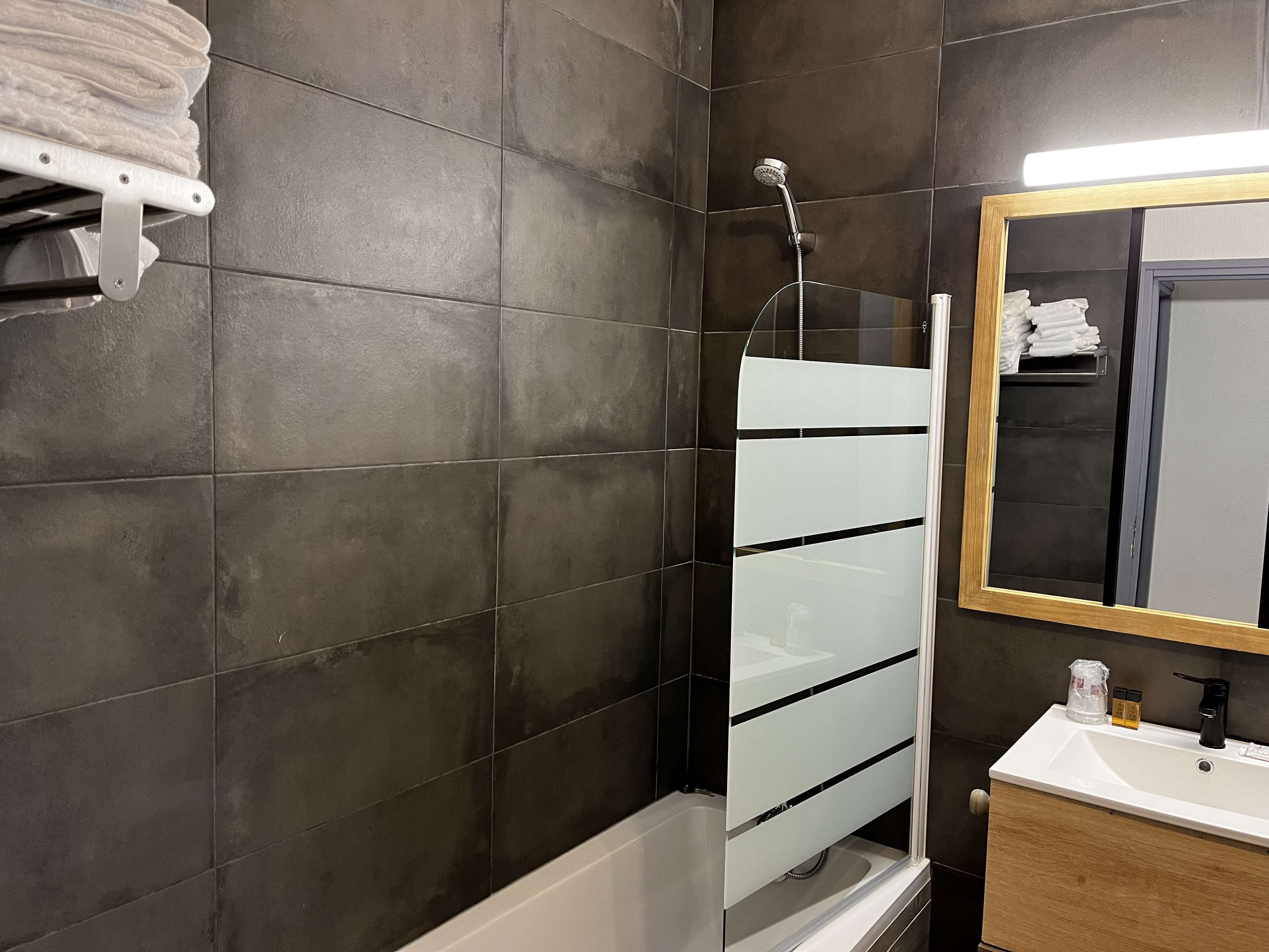 A modern bathroom with dark tiled walls, a glass shower enclosure, a white sink, and a wooden vanity.