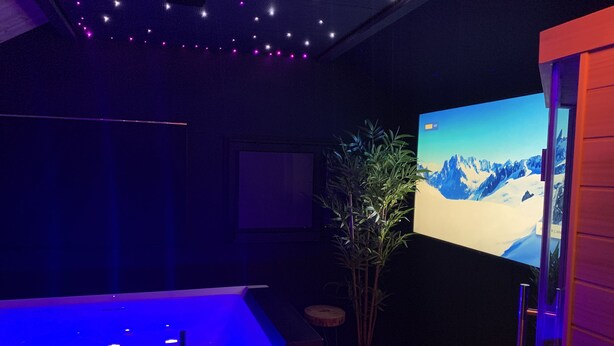 Ein Zimmer mit einem großen Bildschirm, der eine verschneite Berglandschaft zeigt, einer Badewanne mit blauer Beleuchtung und einer Zimmerpflanze.