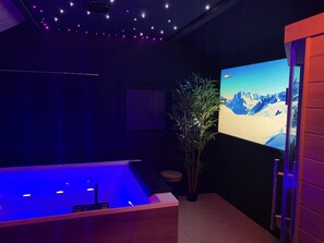 Habitación con una pantalla grande mostrando un paisaje montañoso nevado, una bañera con iluminación azul y una planta en maceta.