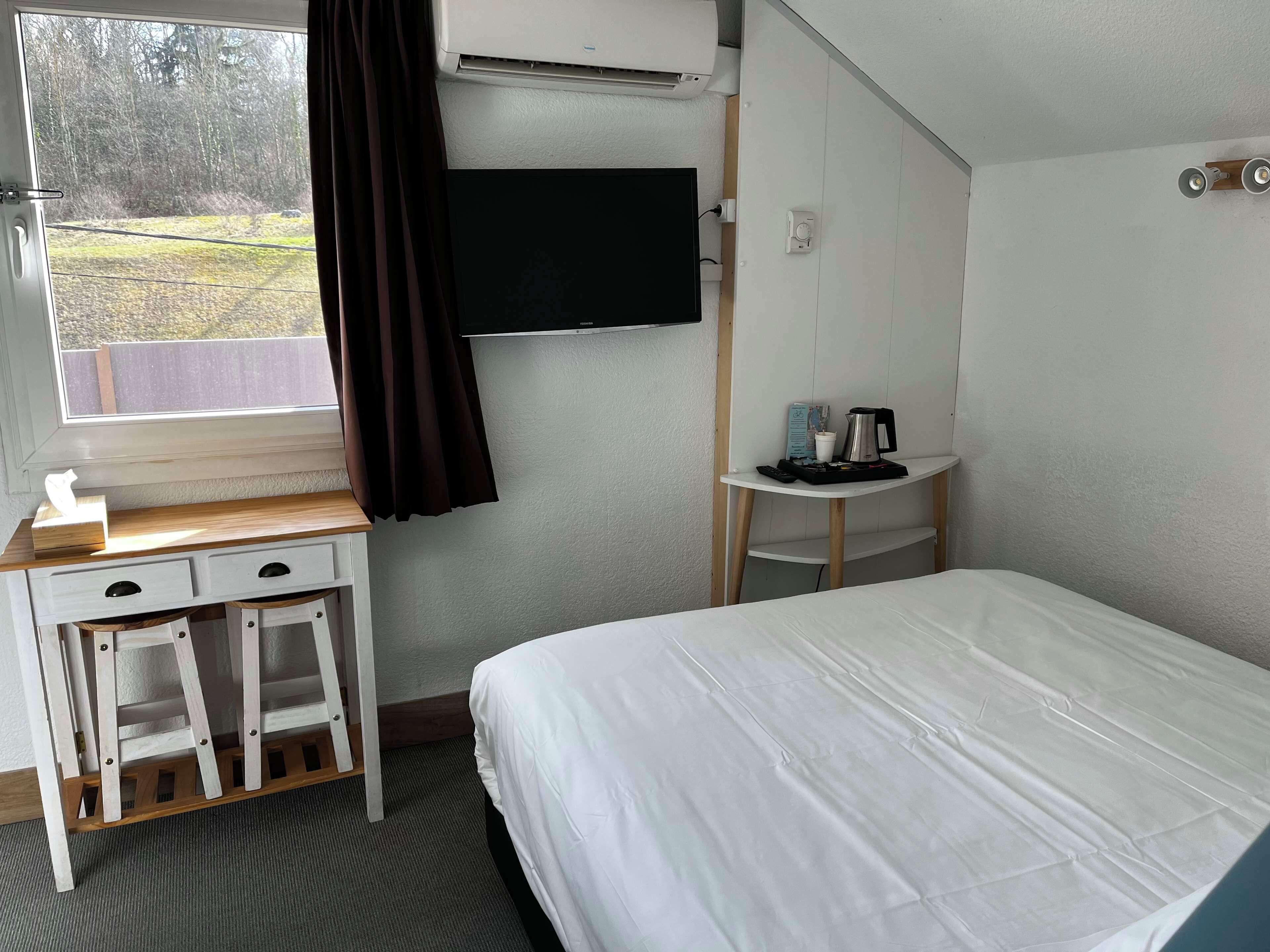 A hotel room with a bed, a desk with drawers, a small table with a coffee maker, a TV mounted on the wall, and a window with curtains.