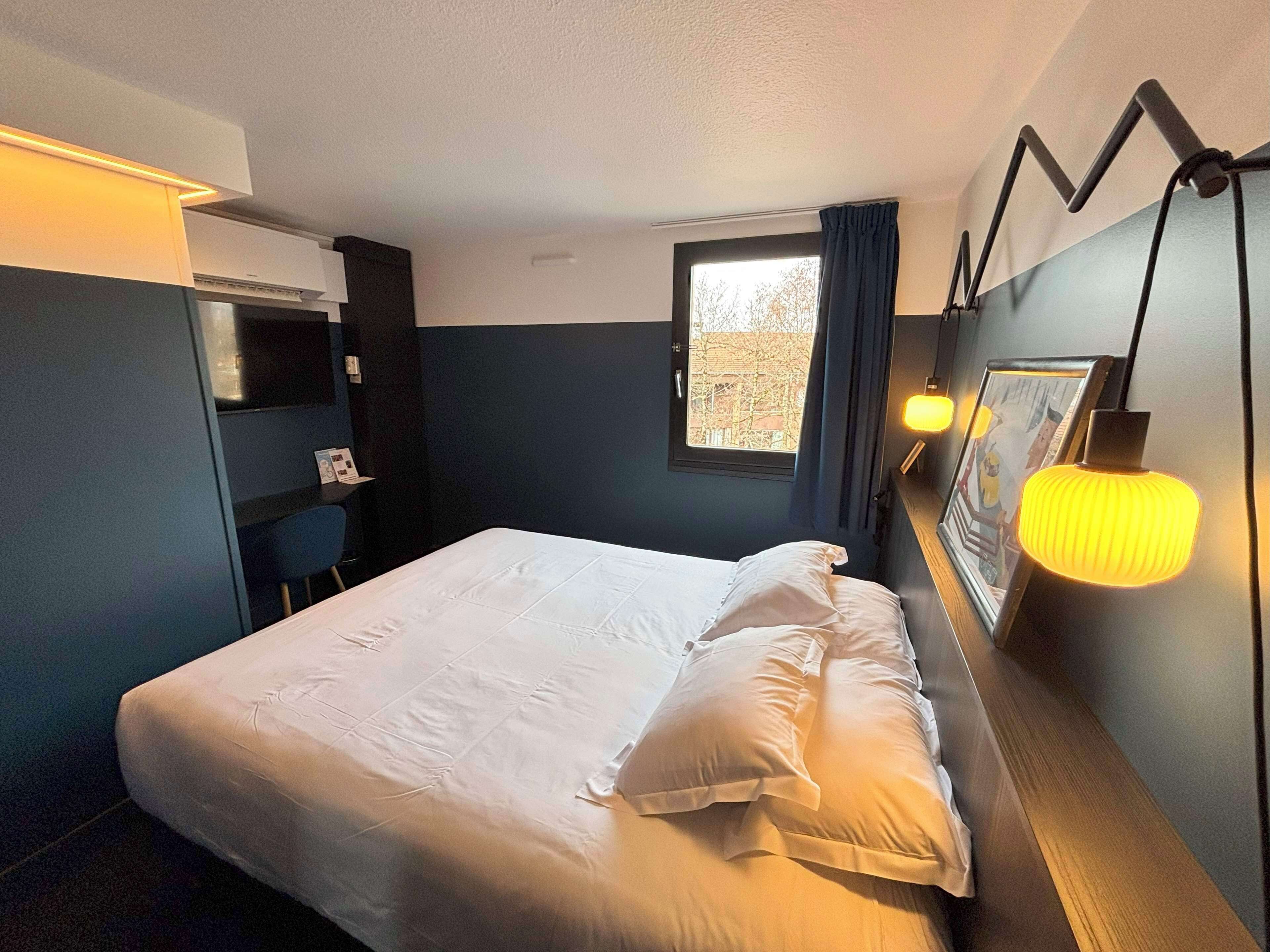 Un lit bien fait dans une chambre d’hôtel, avec une grande fenêtre, un bureau avec une chaise et une lampe fixée au mur.