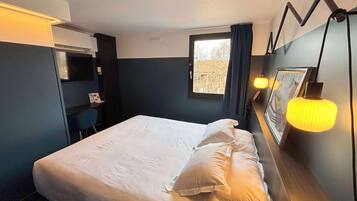 Un lit bien fait dans une chambre d’hôtel, avec une grande fenêtre, un bureau avec une chaise et une lampe fixée au mur.