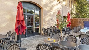Ein Straßencafé mit Korbmöbeln, roten Sonnenschirmen und einem Glastür-Eingang.