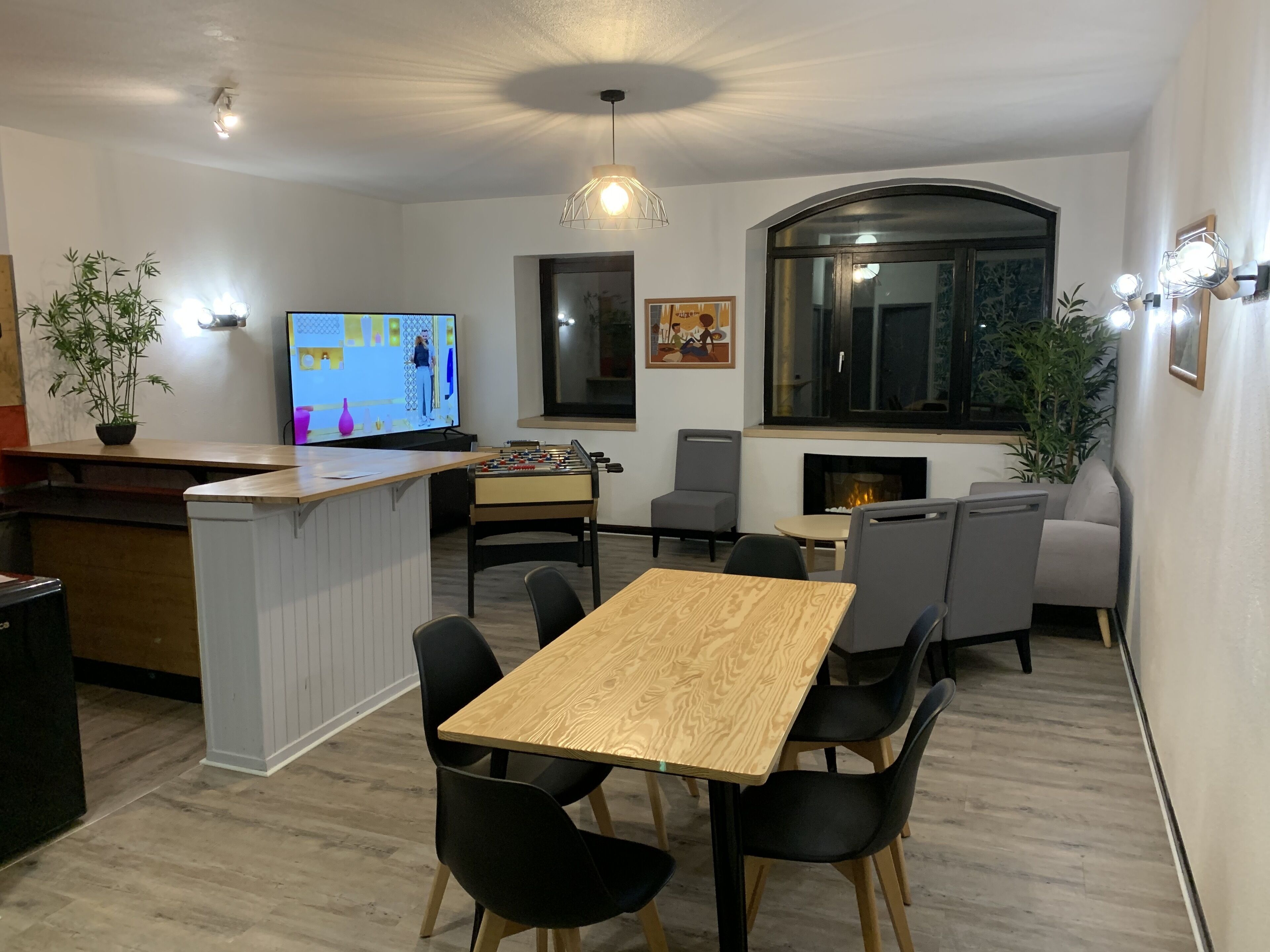 A modern living room with a wooden dining table, black chairs, a foosball table, a TV, and a fireplace.