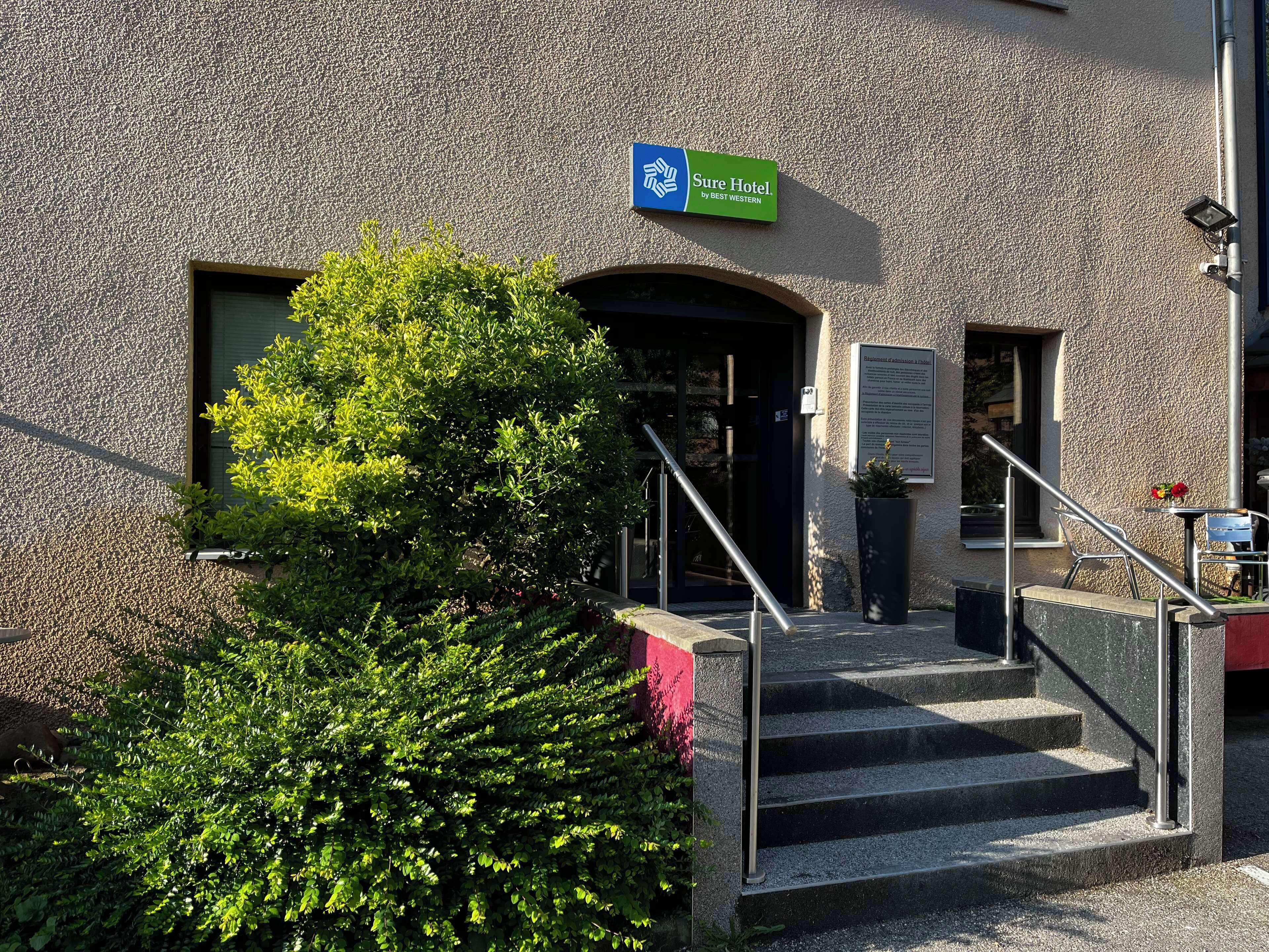 A hotel entrance with a sign reading "Sure Hotel by Best Western," a staircase, and a small outdoor seating area.