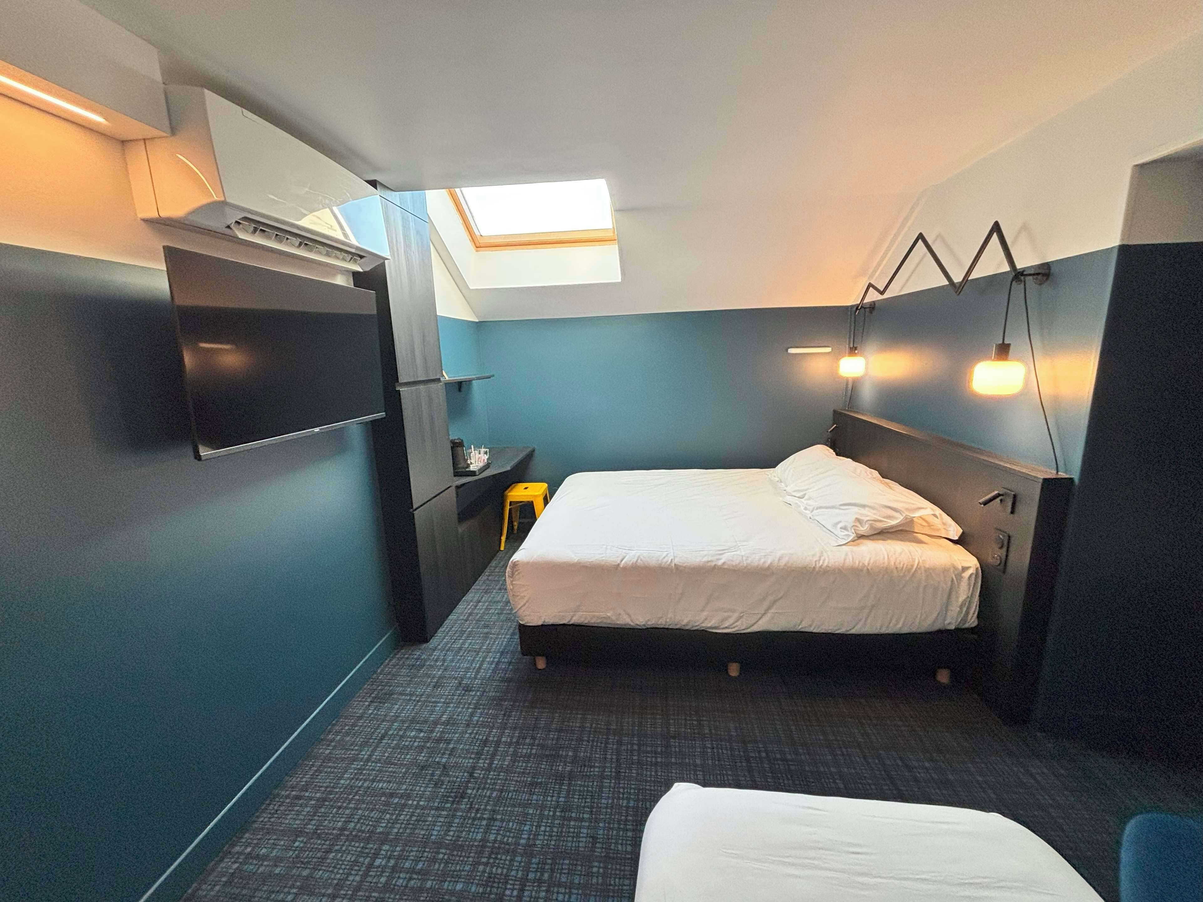 A compact hotel room with a bed, a small desk, and a chair. There is a skylight above the bed and a wall-mounted light fixture.