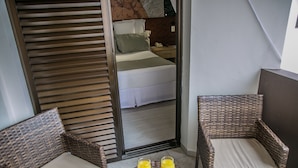 Ein Zimmer mit einem Bett, zwei Korbstühlen, einem kleinen Tisch mit zwei Gläsern Orangensaft und einem Spiegel, der das Zimmer widergibt.