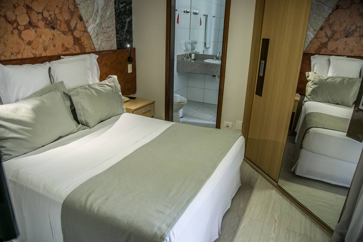 A hotel room with a large bed, a wooden wardrobe, a bathroom with a sink and toilet, and a mirror reflecting the room.