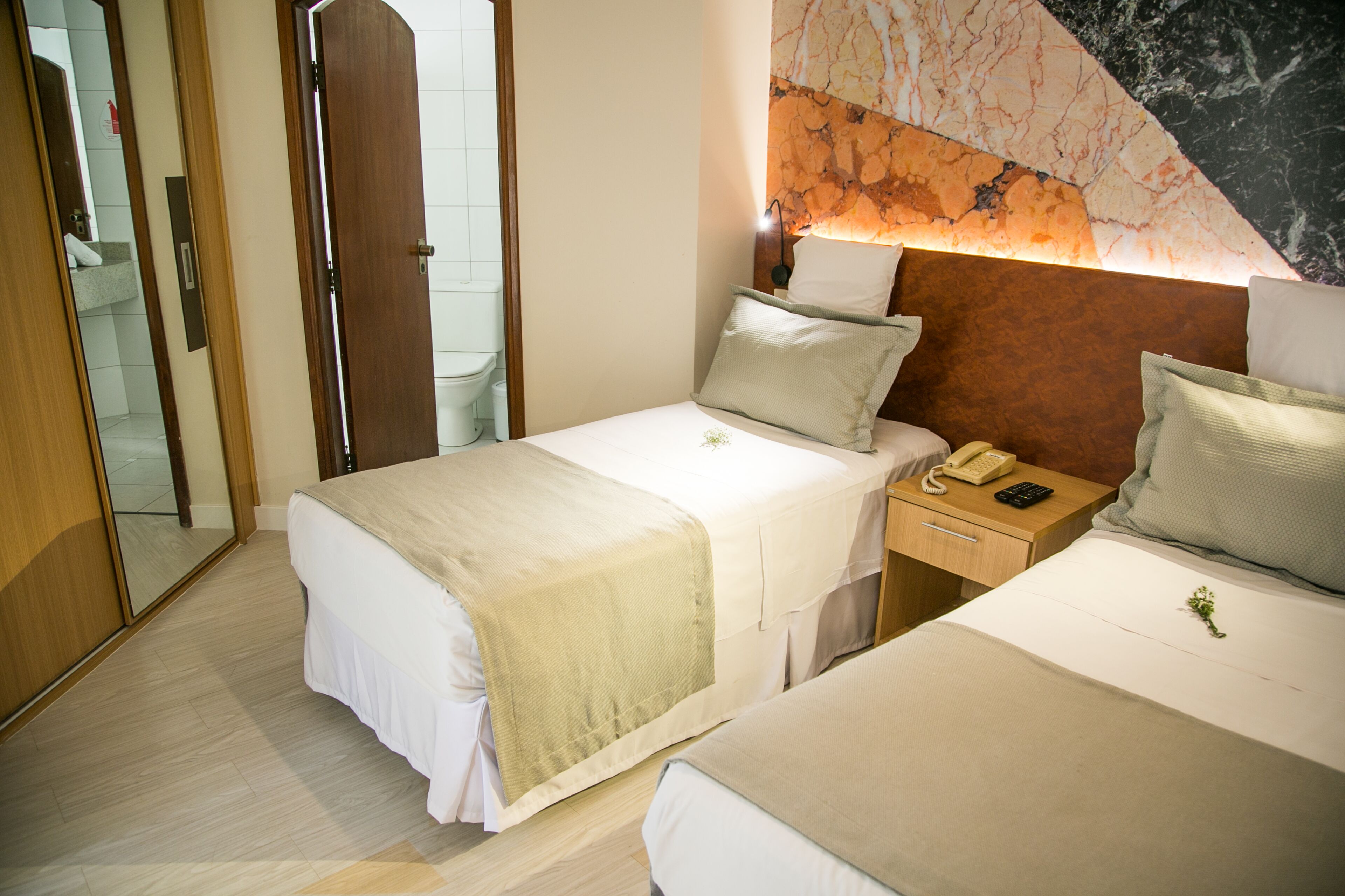 Une chambre d’hôtel avec deux lits, une table de chevet avec un téléphone et une télécommande, et un mur décoré d’un motif imitant le marbre.