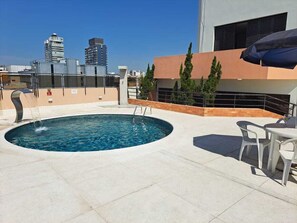 Una piscina en la azotea con vistas despejadas del horizonte urbano, una zona de estar con mesa y sillas, y un edificio moderno con techo plano.