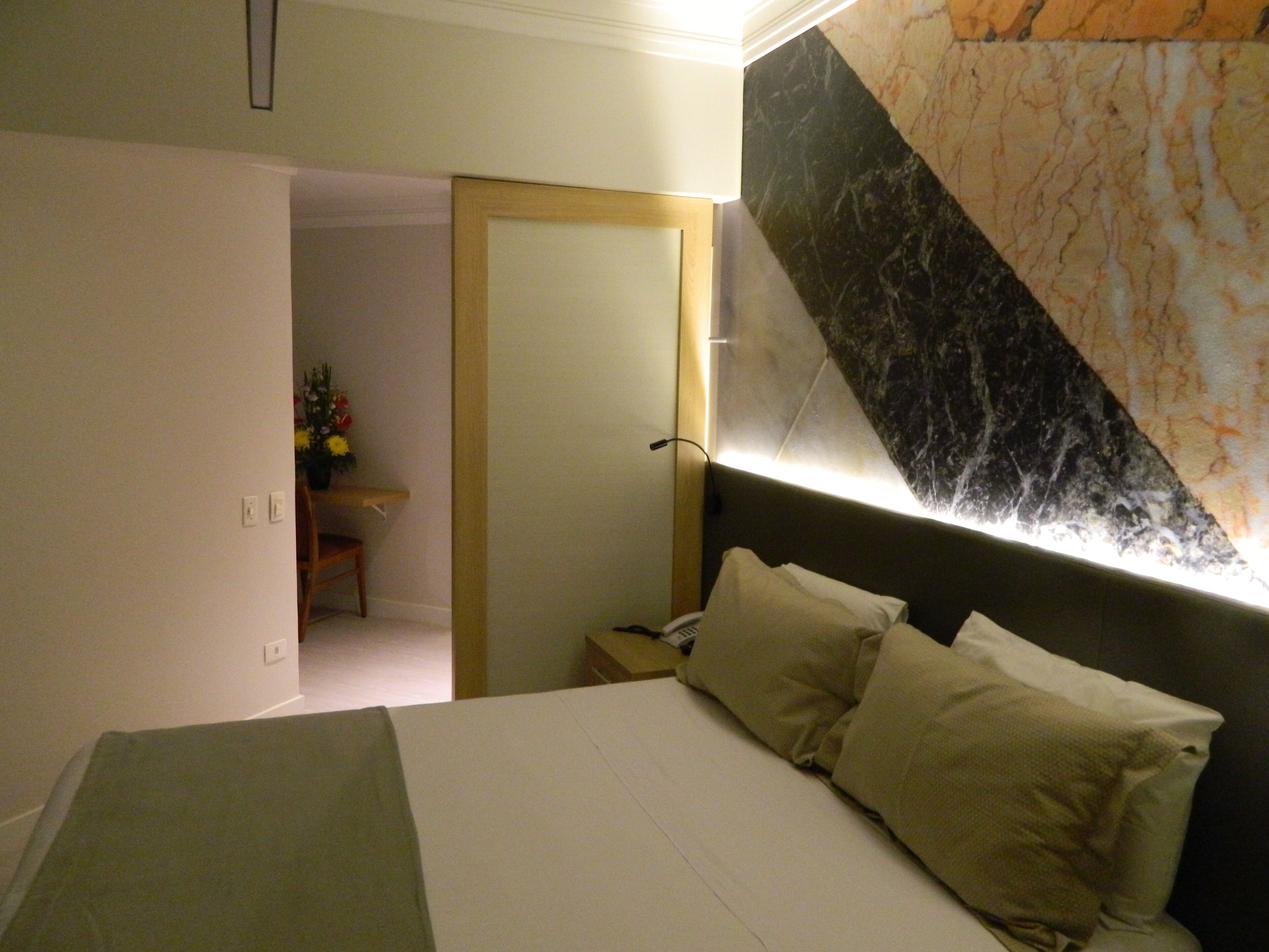 Une chambre à coucher avec un lit, des oreillers, une table de chevet, une lampe, un mur orné d’un motif imitant le marbre et une porte donnant sur une autre pièce.