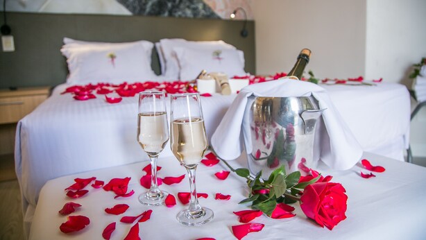 Ein Bett mit weißen Leinentüchern, zwei Champagnergläsern, einer Champagnerflasche und verstreuten Rosenblättern.