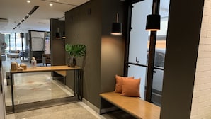 Eine moderne Hotelhalle mit Glastrennwand, einer Holzbank mit Kissen, einer Topfpflanze und Pendelleuchten.