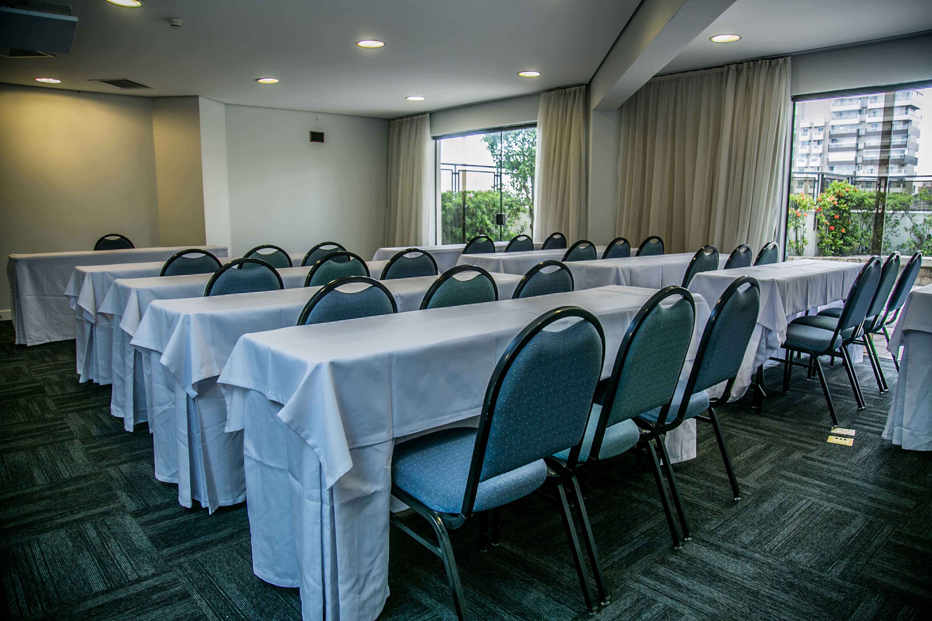 Une salle de conférence dotée de plusieurs tables et chaises, de grandes fenêtres avec des rideaux et d’un sol recouvert de moquette.