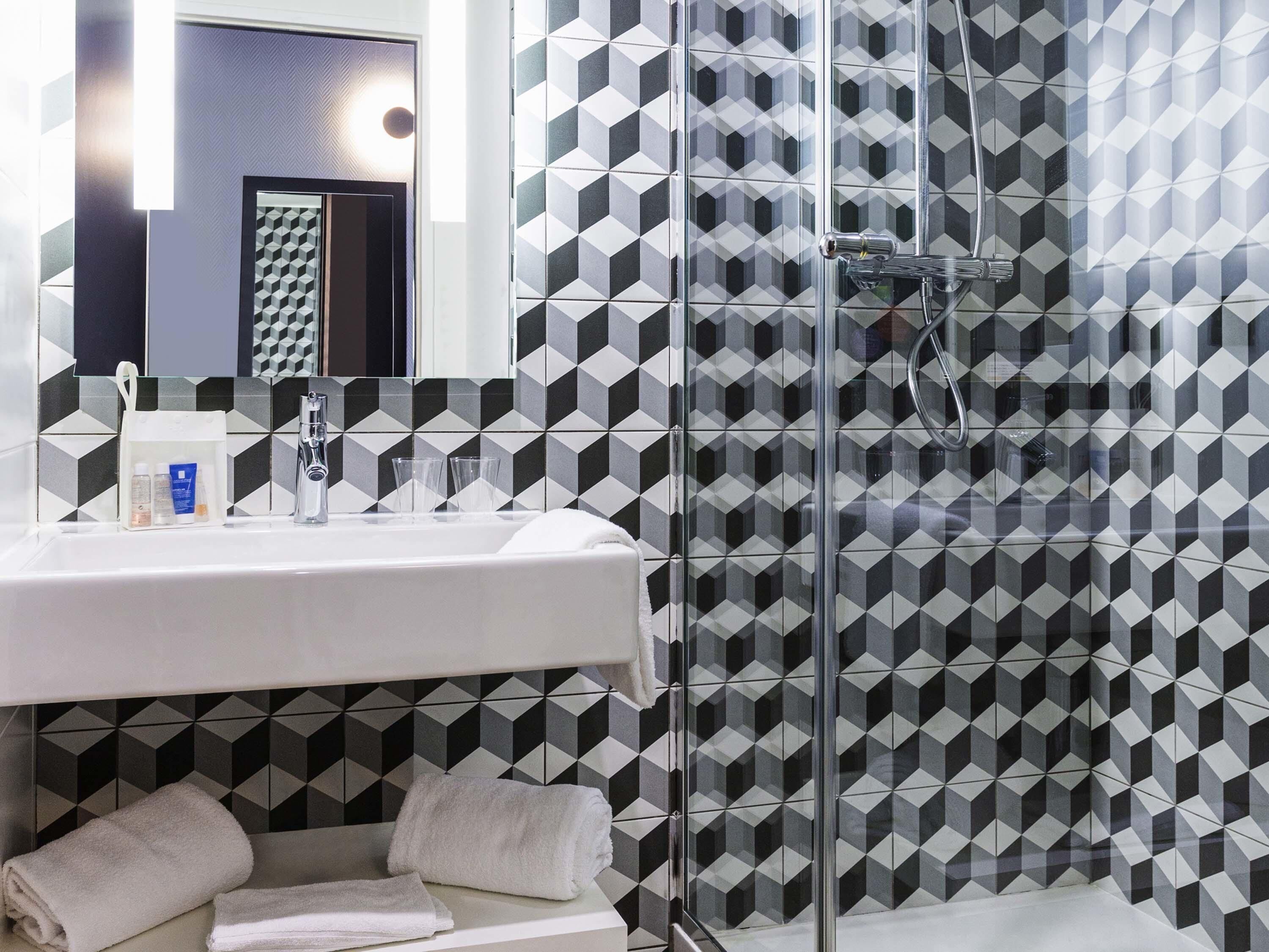 Un baño con pared de diseño geométrico, un lavabo con artículos de aseo personal y una mampara de ducha de vidrio.