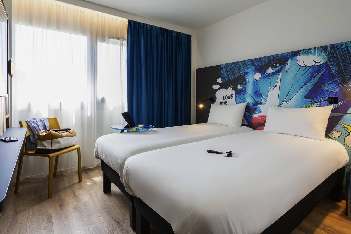 Een hotelkamer met twee bedden, een bureau, een stoel en een groot wanddecor.