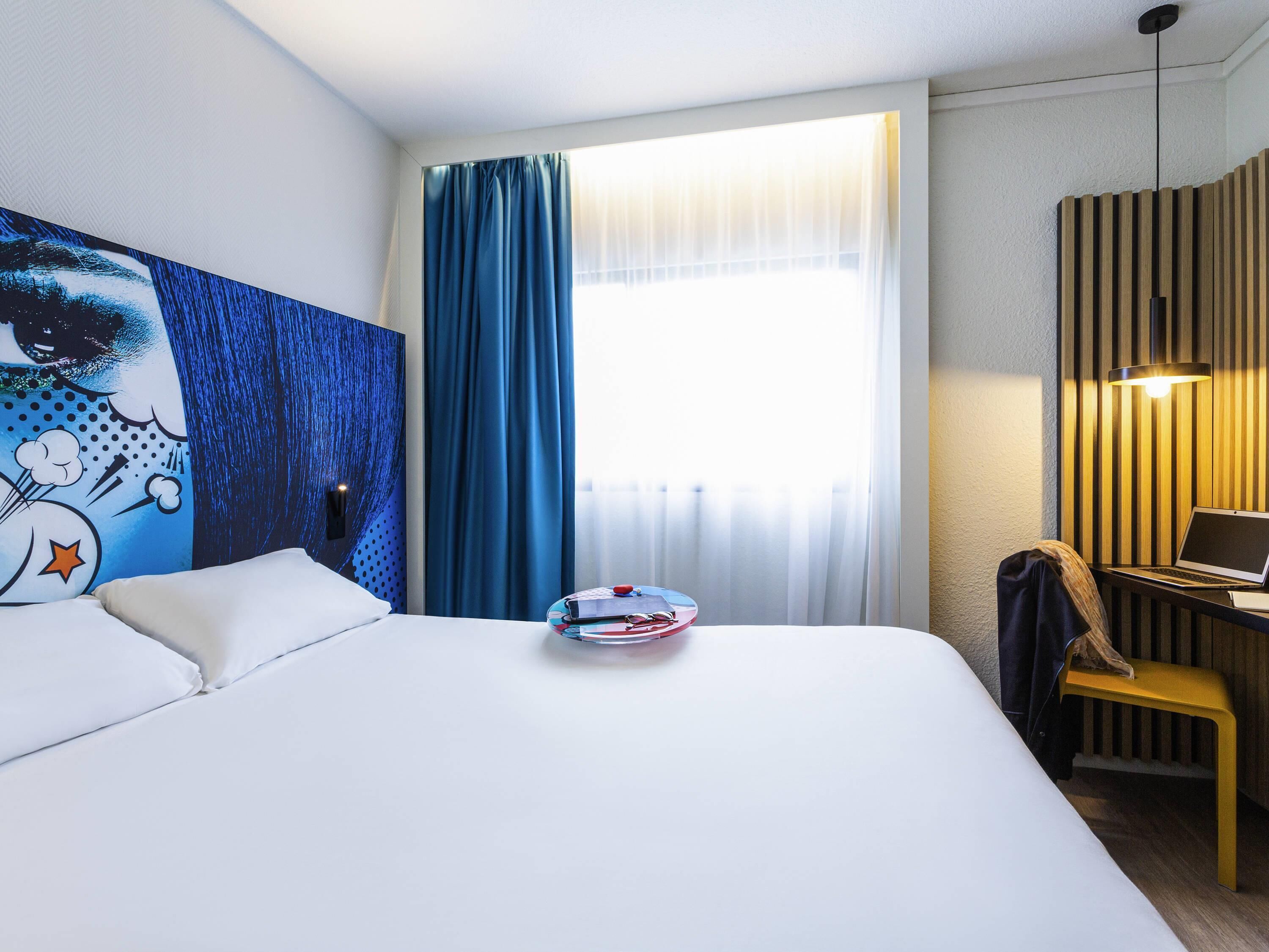 A hotel room with a bed, a desk with a laptop, a painting on the wall, and a window with curtains.