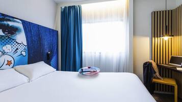 A hotel room with a bed, a desk with a laptop, a painting on the wall, and a window with curtains.