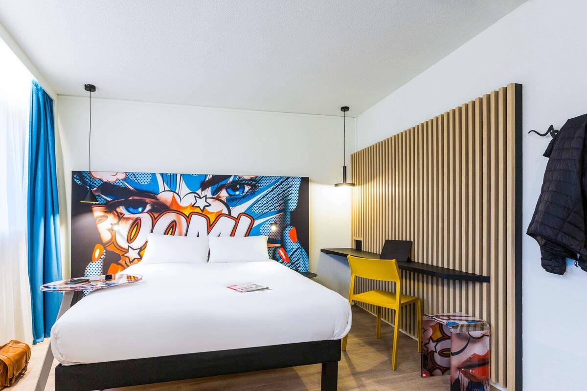 A hotel room with a bed, a desk, a chair, a wall-mounted coat rack, and a colorful artwork on the wall.