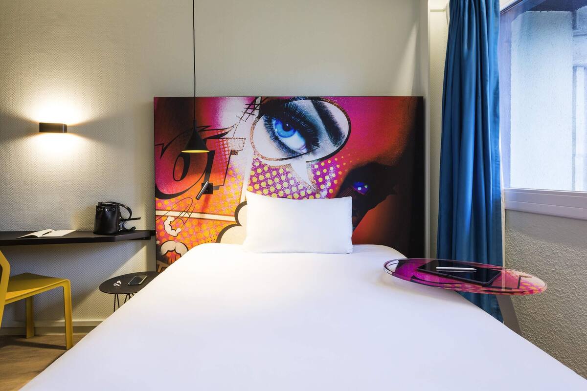 Een hotelkamer met een bed, een bureau met stoel, een tafeltje met telefoon, een wandlamp en een raam met gordijnen.