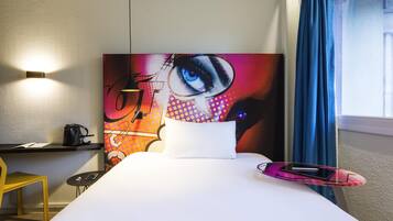 Quarto de hotel com cama, escrivaninha e cadeira, mesinha com telefone, luminária na parede e janela com cortinas.