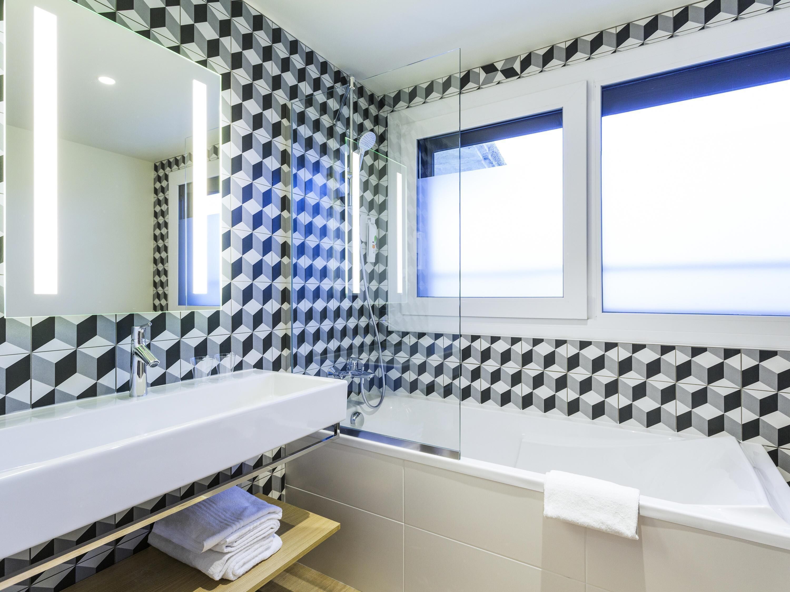 Un baño moderno con un diseño geométrico en los azulejos, un espejo grande, una bañera y una ventana que ofrece vistas.