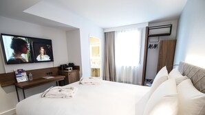 A hotel room with a large bed, a desk with a TV, a minibar, and a bathroom visible through the window.