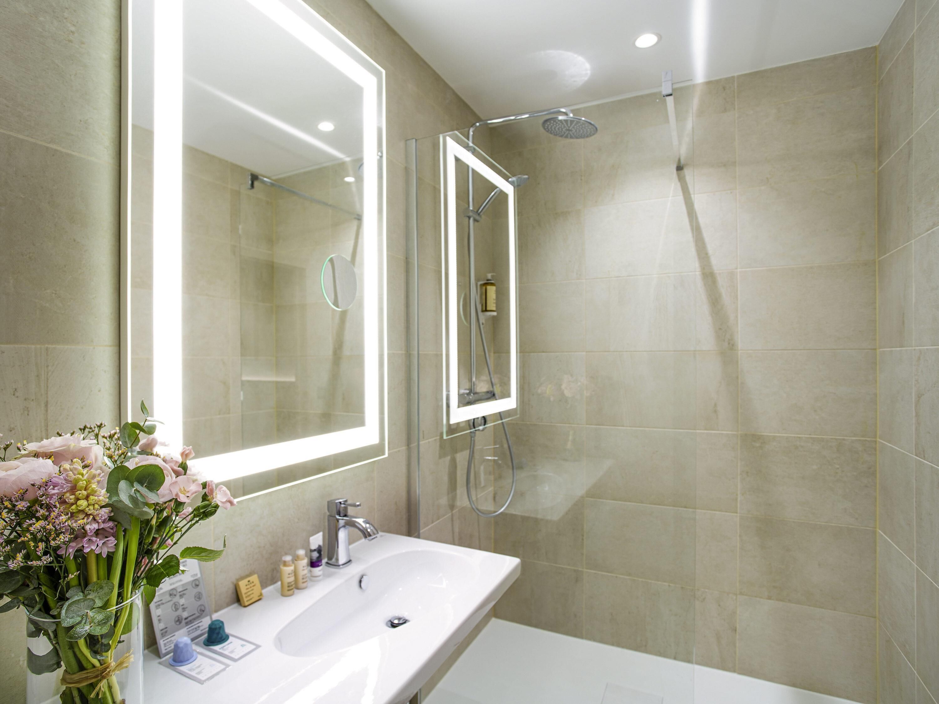 A modern bathroom with a large mirror, a shower, and a sink with toiletries.