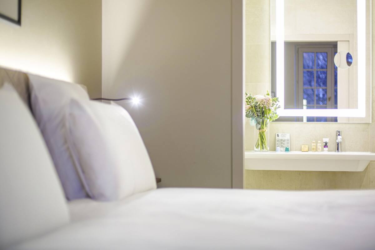 A hotel room with a bed, a bedside lamp, a mirror, a window, and a bathroom counter with toiletries.