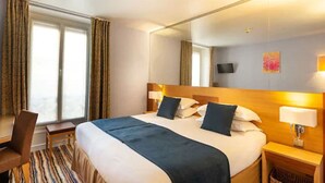 Habitación de hotel con una cama grande, un escritorio, una silla, una ventana con cortinas, una mesita de noche con lámpara y un televisor de pared.