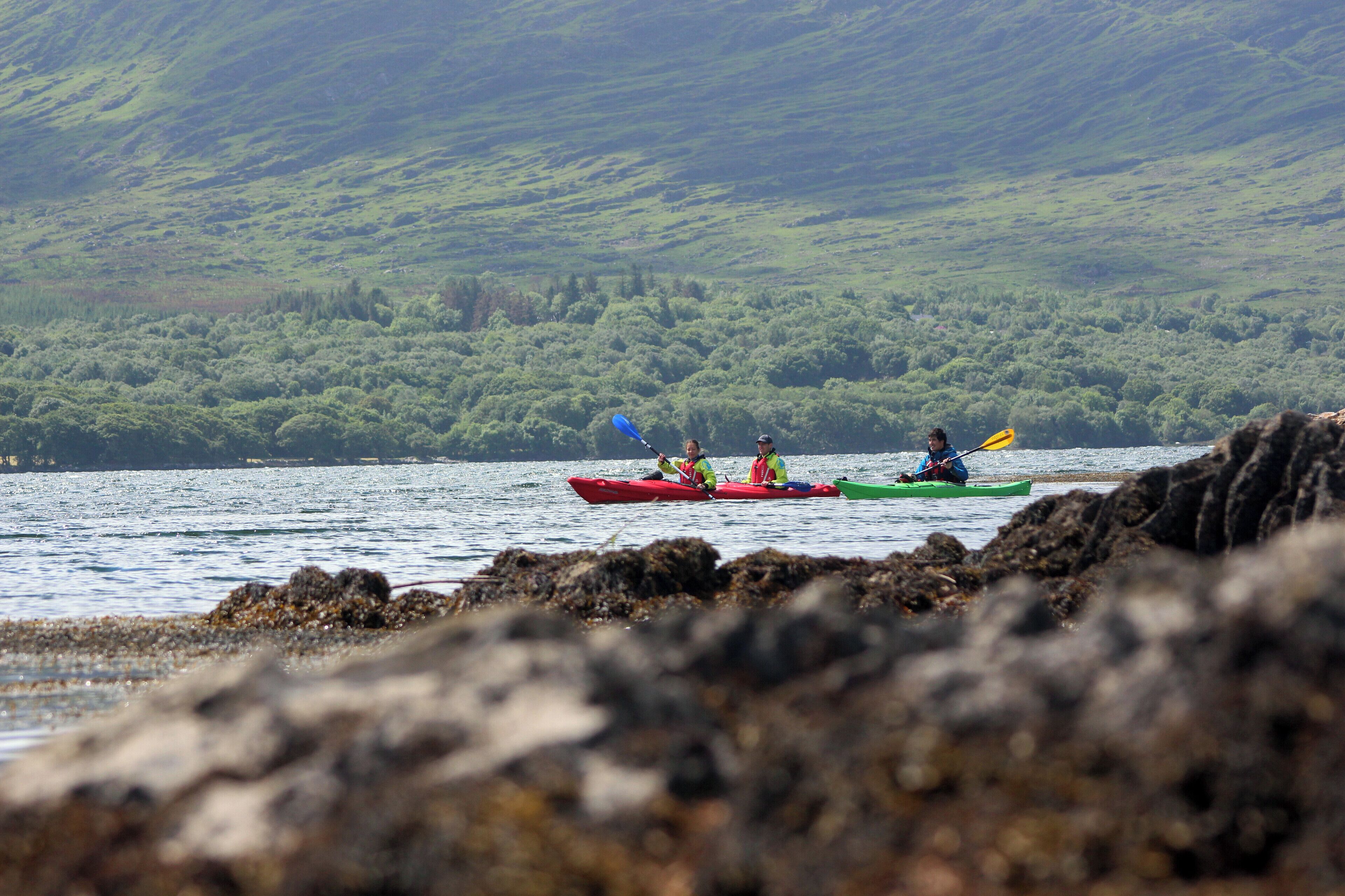 Three kayaks on a calm body of water with a rocky shoreline and a green hillside in the background.