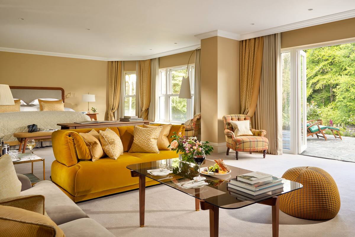 Una sala de estar bien iluminada con un sofá amarillo, una mesa de centro de vidrio y un sillón cómodo. Se ve un ventanal con vistas al exterior.