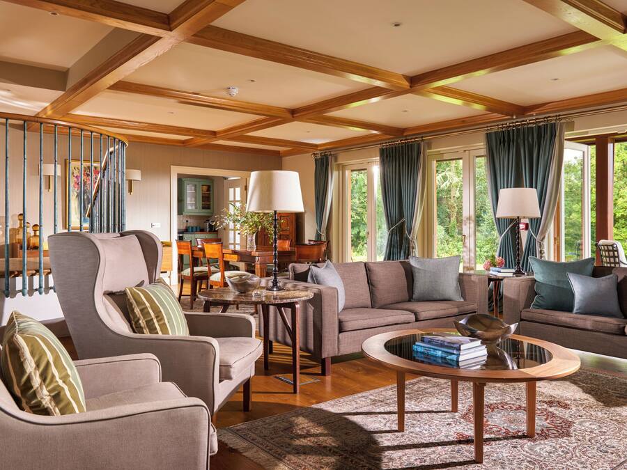 A spacious living room with a high ceiling, wooden beams, a dining area, and large windows with curtains.