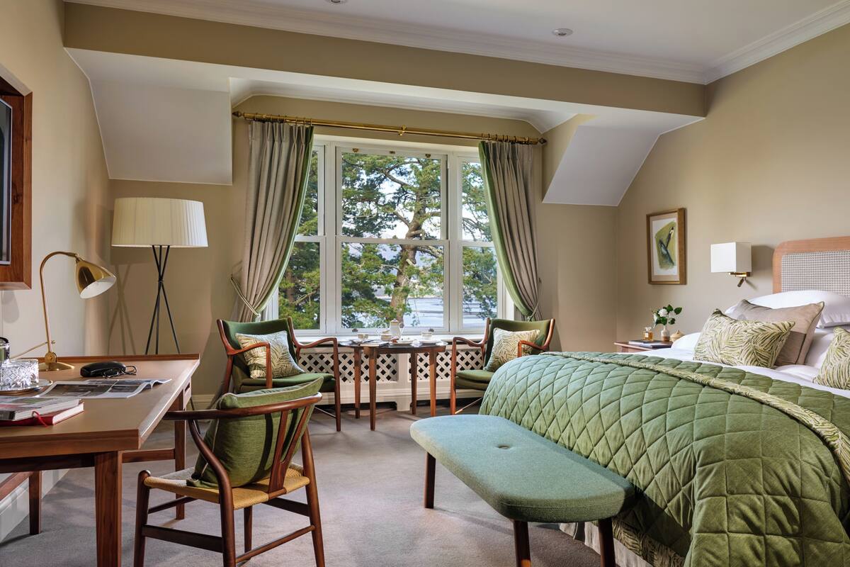 Habitación de hotel con cama, escritorio, sillas, ventana con cortinas y vista a árboles.