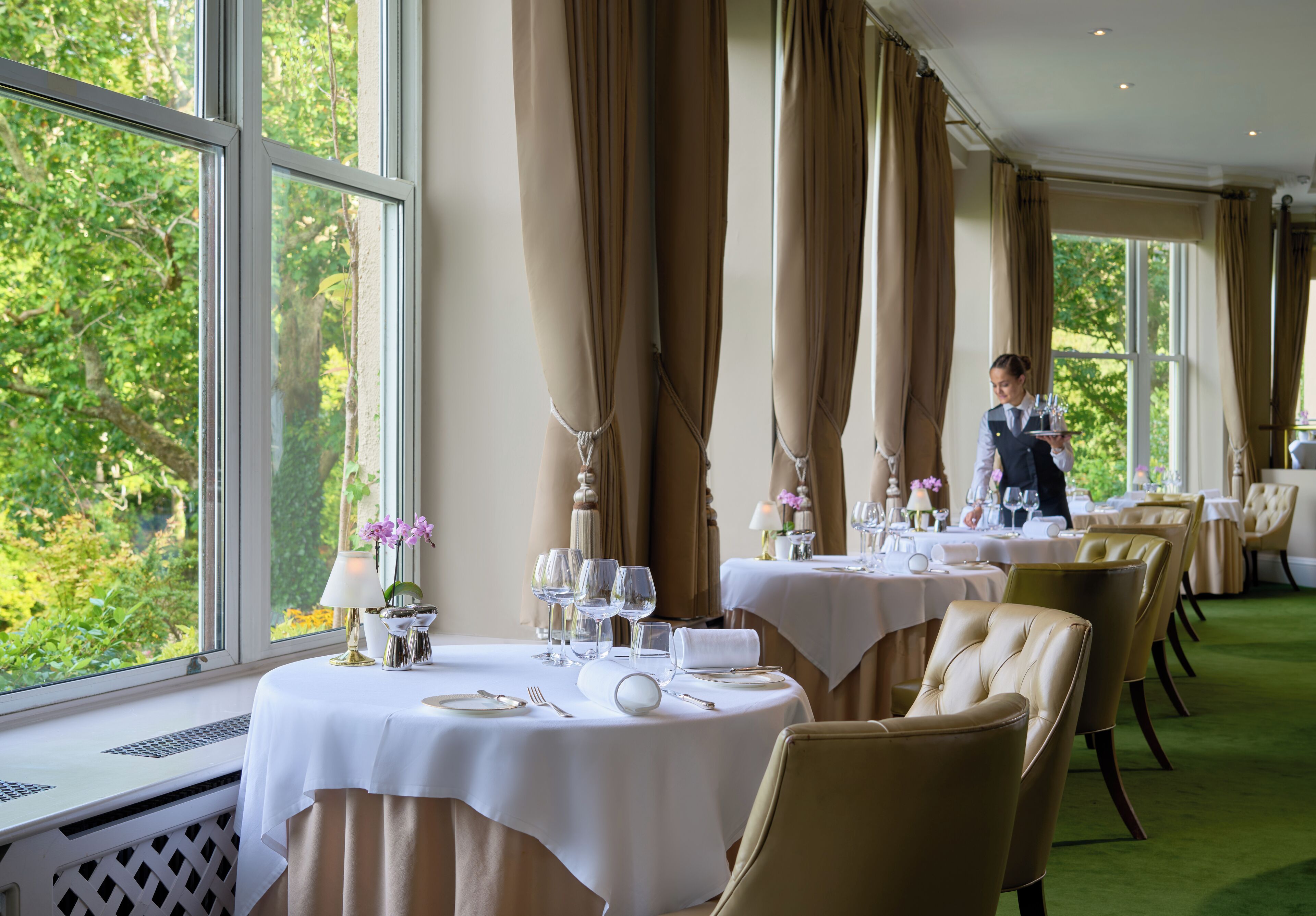 Un restaurant avec des tables dressées pour le service, de grandes fenêtres ornées de rideaux et une vue sur la verdure environnante.