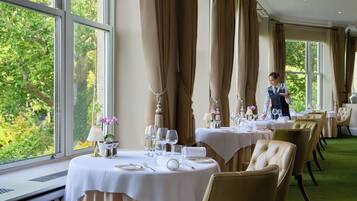 Een restaurant met gedekte tafels, grote ramen met gordijnen en uitzicht op groen.
