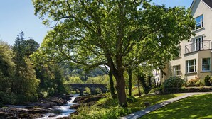 Un río serpentea por un parque con un puente de piedra, rodeado de abundante vegetación y un edificio con balcón.